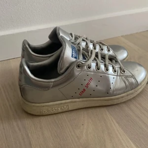 Silvriga Adidas Stan Smith sneakers - Snygga Adidas Stan Smith sneakers i silver med klassisk blå logga på plösen och röd text på sidan. Skorna har snörning, rund tå och platt sula.  Perfekta för dig som vill sticka ut med en ikonisk sneaker.