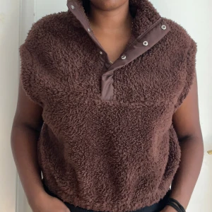 Brun ärmlös fleece-tröja med krage - Mysig brun ärmlös fleece-tröja med hög krage och tryckknappar framtill. Perfekt för lager-på-lager och ger en cool, avslappnad vibe. Tröjan har en loose passform och är supermjuk mot huden.