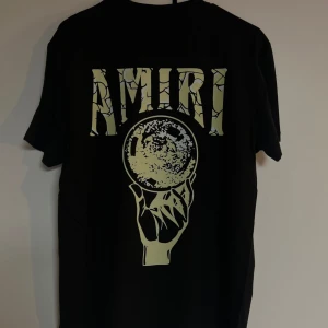 Svart Amiri t-shirt med tryck - Svart t-shirt från Amiri med stort grafiskt tryck på ryggen som visar en hand som håller en klot och texten 'AMIRI' i sprucken stil. Framsidan har en färgglad droppande logga. Skön passform och tillverkad i mjuk bomull.