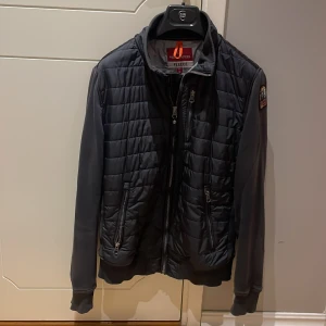 Svart kardigan/jacka Parajumpers - Snygg svart quiltad jacka från Parajumpers med broderad patch på ärmen. Jackan har dragkedja framtill, två dragkedjefickor och ribbade muddar. Jackan är ett mellanting mellan jacka och tröja. Ärmar i mjuk bomull och fram- och bakstycke vadderat. Storlek small, passar kille som tjej.