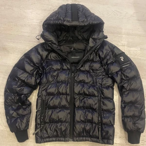 Svart pufferjacka Peak Performance S - Snygg svart pufferjacka från Peak Performance i storlek S. Jackan har huva, dragkedja framtill och två sidofickor med dragkedja. Perfekt för kalla dagar med sin quiltade design och sportiga look. Klassisk logga på ärmen och sköna muddar vid ärmsluten.