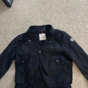 Moncler windbreaker - Säljer en svart Moncler Donatien vindjacka med klassisk logga på ärmen. Jackan har flera dragkedjeförsedda fickor, hög krage och stilren design. Perfekt för dig som vill ha en snygg och funktionell jacka med exklusiv känsla.