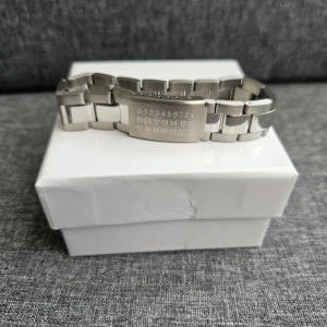 Maison Margiela silver armband - Coolt armband från Maison Margiela i blankt silverfärgat metall med deras ikoniska siffror ingraverade på plattan. Länkarna är chunky och ger en modern vibe, perfekt för dig som gillar statement-accessoarer. Snyggt att bära till både streetwear och mer dressade outfits.