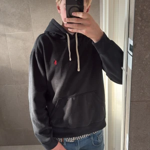 Svart hoodie från Polo Ralph Lauren - Svart hoodie från Polo Ralph Lauren med vit snörning och röd broderad logga på bröstet. Klassisk känguruficka framtill och huva. Skön och stilren modell i bomull, perfekt till jeans eller joggers.