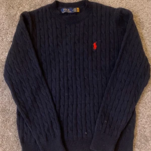 Mörkblå stickad tröja från Polo Ralph Lauren - !Helt oanvänd !köp i Paris Säljer en klassisk mörkblå stickad tröja från Polo Ralph Lauren med rund halsringning och röd broderad logga på bröstet. Tröjan har kabelstickat mönster Perfekt för en stilren och tidlös look.