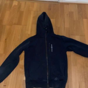 Sail racing hoodie - Svart hoodie med dragkedja från Sail racing. Klassisk modell med huva och fickor framtill. Perfekt för både träning och vardag. Diskret logga på bröstet och mjukt material.