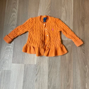 Orange kofta från Ralph Lauren, 24M - Superfin orange kofta från Ralph Lauren med kabelstickat mönster, volangkant nedtill och pärlemorknappar. Tröjan är i 100% bomull och har långa ärmar samt en liten rosa logga broderad på bröstet. Perfekt för att liva upp din lillas outfit! Fåtals gånger använd!
