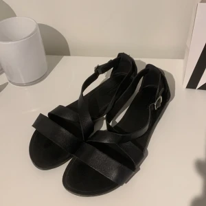 Svarta sandaler i skinn med spänne - Stilrena svarta sandaler med remmar i skinn och justerbart spänne vid ankeln. Liten klack. Snyggt korsade remmar framtill och silverfärgat spänne på sidan.