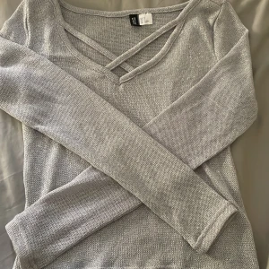 Ljusgrå glitter stickad tröja med v-ringning från H&M Divided - Säljer en ljusgrå stickad tröja från H&M Divided i storlek XS. Tröjan har v-ringning med dekorativa korsade band framtill och långa ärmar. Perfekt för lager-på-lager och enkel att matcha med olika outfits.