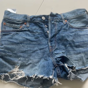 Blå jeansshorts  från Levi's - Säljer ett par klassiska blå jeansshorts från Levi's med råa fransade kanter och knappgylf. Shortsen har fyra fickor och en avslappnad passform, perfekt för varma dagar.