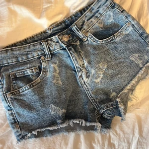 Blå jeansshorts med fransar - Säljer ett par blå jeansshorts med slitna detaljer och fransig kant nedtill. Klassisk femficksmodell med knapp och dragkedja framtill. Perfekta för sommaren och ger en avslappnad vibe. Storlek 36