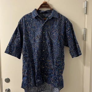 Blå mönstrad skjorta från Colours by Alexander Julian - Säljer en blå kortärmad skjorta från Colours by Alexander Julian. Skjortan har ett färgstarkt abstrakt mönster i vitt, orange och gult samt knappar framtill. Tillverkad i 100% bomull, perfekt för varma dagar.