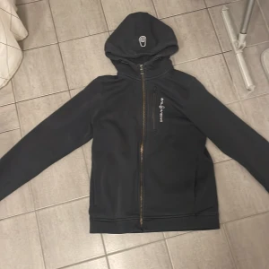 Svart vindjacka från Sail Racing - Svart vindjacka från Sail Racing med huva och dragkedja framtill. Jackan har en bröstficka med dragkedja och logga broderad på bröstet samt på huvan. Snygg och sportig modell som passar perfekt till vardagsoutfiten.