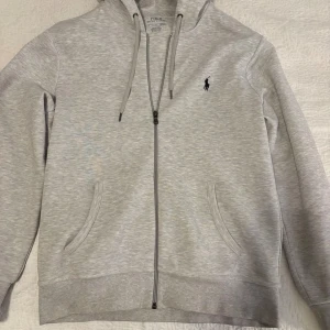  Polo Ralph Lauren - Säljer en stilren ljusgrå hoodie från Polo Ralph Lauren med dragkedja och huva. Tröjan har två fickor framtill och den klassiska broderade loggan på bröstet. Perfekt för en avslappnad look.