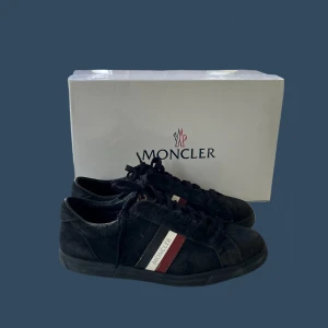 Moncler sneakers - Snygga svarta sneakers från Moncler med röd och vit randdetalj på sidan. Skorna har snörning och är perfekta för en stilren look. Levereras med originalförpackning och dustbags. ALLT OG INGÅR! Kom med bud