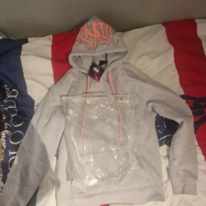 Ljusgrå hoodie från syna world med rosa detaljer - Säljer en ljusgrå hoodie från syna world med rosa dragsnören och tryck på huvan. Tröjan har en stor magficka och är perfekt för en avslappnad stil. Mjuk och skön, passar till många tillfällen.