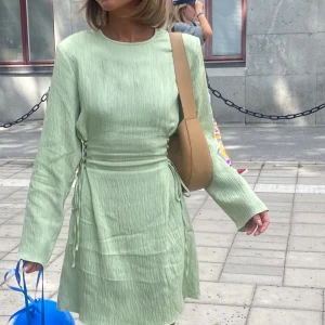 Ljusgrön omlottklänning från MAUD - Ljusgrön kortklänning ”Amelia dress” från märket MAUD med lång ärm, rund halsringning och små axelvaddar. Använd endast en gång och är i nyskick. Köptes för 1400 kr. Klänningen har snygga knyt- och omlottdetaljer i midjan samt en lätt skrynklad struktur i tyget. Perfekt för sommarfesten! 