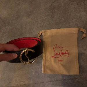 Svart bälte med guldfärgat spänne från Christian Louboutin - Säljer ett svart bälte från Christian Louboutin med röd insida och ett stort, guldfärgat spänne i form av CL-loggan. Bältet är tillverkat i skinn och har en exklusiv känsla med tydliga detaljer.