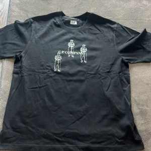 Svart t-shirt från C.P. Company - Säljer en svart t-shirt från C.P. Company med tryck av sittande figurer och logga på både fram- och baksida. Klassisk rund hals och korta ärmar. Perfekt för dig som gillar streetwear och vill ha något stilrent men ändå unikt. 