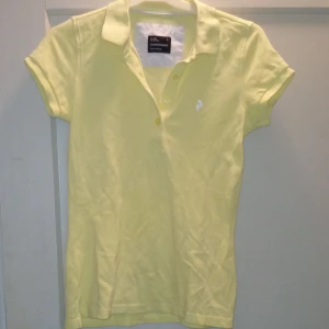 Gul pikétröja från Peak Performance - Säljer en ljusgul pikétröja från Peak Performance med klassisk krage och korta ärmar. Tröjan har knäppning framtill och en liten vit logga broderad på bröstet. Perfekt för en sportig och avslappnad stil.