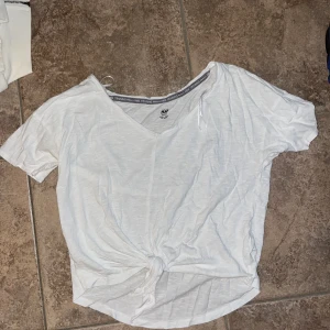 Vit knyt-topp från H&M Training - Vit kortärmad topp från H&M Training med v-ringning och snygg knytning framtill. Tunt och lätt material som passar perfekt till träning eller chill. Enkel och stilren design som är lätt att matcha med andra plagg.