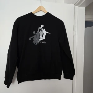 Svart sweatshirt med baskettryck H&M - Svart sweatshirt från H&M med coolt basketmotiv i grått och vitt på bröstet. Tröjan har rund hals, långa ärmar och är gjord i mjuk bomullsblandning med lite polyester. Perfekt för dig som gillar sportig stil och streetwear. Katt finns I hemmet