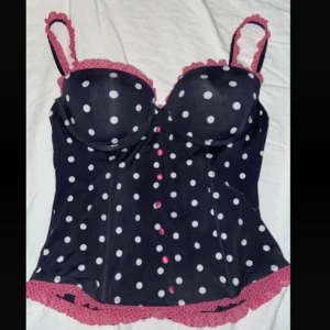 Svart prickig korsett med rosa spets - Supersöt svart korsett med vita prickar och rosa spetsdetaljer längs kanterna. Axelbanden är dekorerade med rosa volang och framtill finns små rosa knappar. Cupan är i storlek 36C💞💞
