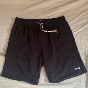 Svarta shorts från BOSS med snörning - Svarta shorts från BOSS med vit logga på vänster ben och justerbar snörning i midjan. Klassisk och stilren design, perfekt för avslappnade dagar eller träning. Mjukt material och bekväm passform.