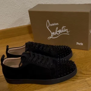 Svarta sneakers med nitar från Christian Louboutin - Säljer ett par svarta sneakers från Christian Louboutin med svarta nitar på tån och sidorna. Skorna har snörning och en platt sula. Perfekta för dig som vill sticka ut med en lyxig och edgy stil.