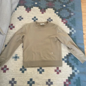 Beige sweatshirt från Acne Studios - Säljer en stilren beige sweatshirt från Acne Studios med broderade bokstäver 'A S' på framsidan. Tröjan har rund halsringning och långa ärmar. Perfekt för en clean och avslappnad look.