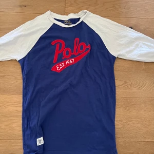 Blå och vit tröja från Polo Ralph Lauren - Säljer en blå tröja med vita ärmar från Polo Ralph Lauren. Tröjan har röd broderad text 'Polo EST. 1967' på bröstet och klassisk basebollstil med raglanärmar. Perfekt för en sportig look!