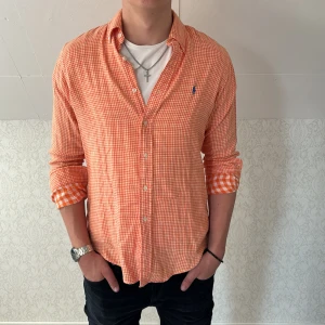 Orange-rutig skjorta från Polo Ralph Lauren - Säljer en orange och vit rutig skjorta från Polo Ralph Lauren i modellen Custom Fit. Skjortan har långa ärmar, klassisk krage och blå broderad logga på bröstet. Perfekt för dig som gillar färg och stilren design.