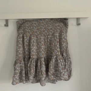 Blommig volangkjol från 157 - Supersöt kort blommig kjol från 157 i beige och vitt. Kjolen har smock i midjan och volang nertill, vilket ger en luftig och skön känsla. Perfekt för varma dagar och enkel att styla med en enkel topp.