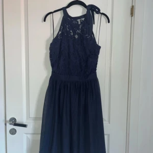 Marinblå spetsklänning från Vila - Säljer en marinblå klänning från Vila med spetsdetaljer upptill och knytning vid axeln. Klänningen har ärmlös design och en lätt utställd kjol i chiffongliknande material. Perfekt för speciella tillfällen.  Strl 38