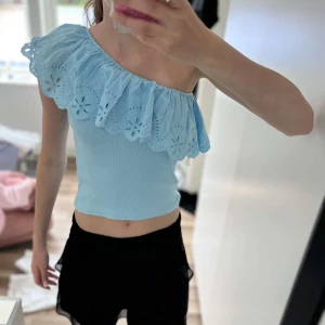 Ljusblå one shoulder topp med volang från Zara - Superfin ljusblå topp från Zara med one shoulder-design och bred volang med broderade blommor. Toppen har en ribbad struktur i tyget. Perfekt för varma dagar och ger en söt och trendig look. Säljer för att den är för liten! Priset går att diskuteras!