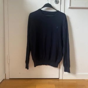 Mörkblå stickad tröja från Ralph Lauren - Mörkblå stickad tröja från Ralph Lauren med rund halsringning och diskret broderad logga på bröstet. Tröjan har långa ärmar och ribbade muddar vid ärmslut och nederkant. Perfekt för lager-på-lager-stil. Helt ok skick skriv vid frågor