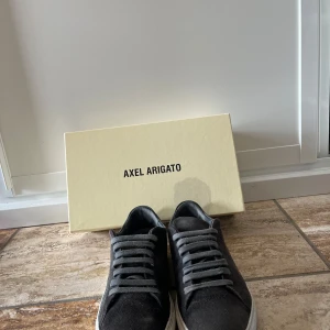 Gråa sneakers från Axel Arigato - Säljer ett par stilrena gråa sneakers från Axel Arigato med ovandel i mocka och vit platt sula. Skorna har rund tå och klassisk snörning. Perfekta för dig som gillar minimalistisk design och hög kvalitet. Medföljer påse. Hör av er om ni har några frågor.