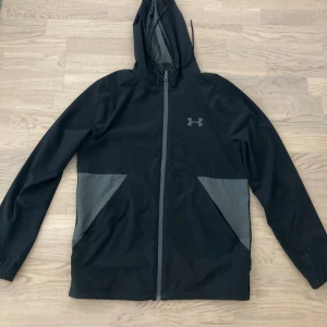 Svart vindjacka från Under Armour med huva - Säljer en svart vindjacka från Under Armour med grå detaljer och huva. Jackan har dragkedja framtill, två stora fickor med grått mesh-tyg och justerbar dragsko i huvan. Perfekt för träning eller utomhusaktiviteter.