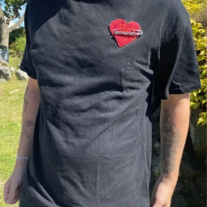 Svart t-shirt med rött hjärta från Palm Angels - Säljer en svart t-shirt från Palm Angels med ett broderat rött hjärta och säkerhetsnål-detalj på bröstet. Klassisk passform och rund halsringning. Perfekt för dig som gillar streetwear och vill sticka ut lite extra.