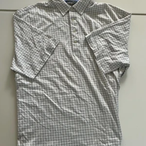 Rutig stjortpiké från Polo Golf Ralph Lauren - Snygg rutig henley-skjorta från Polo Golf Ralph Lauren i ljusgrått och vitt. Skjortan har klassisk krage, korta ärmar och knappar framtill. Tillverkad i mjuk bomull, perfekt för en avslappnad men stilren look.