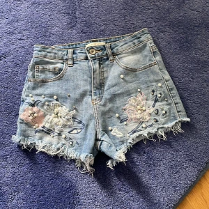 Blommiga jeansshorts från Miss Bonbon - Snygga ljusblå jeansshorts från Miss Bonbon. Fransig kant och broderade blommor samt pärldetaljer framtill. Perfekta för sommaren och ger en cool, trendig vibe. Materialmix med bomull, viskos och polyester för skön passform.