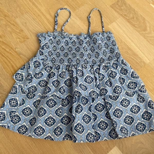 Blå mönstrad topp Gina Tricot - Supersöt blå och vit topp från Gina Tricot med smala axelband och smock upptill. Toppen har ett snyggt mönster i mörkblått och vitt och är perfekt för varma dagar. Lätt och luftig känsla, passar till både jeans och shorts.