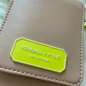 Beige axelväska från Kendall + Kylie -  beige axelväska från Kendall + Kylie med neon-gula detaljer och axelrem. Väskan har ett fack med magnetknapp, kortfack på insidan och transparent baksida med logga. Tillverkad i syntetmaterial och polyester.
