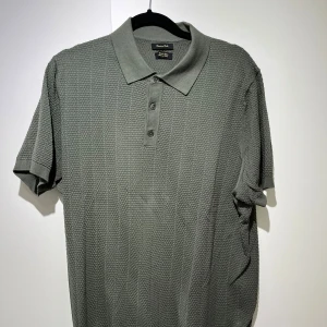 Grön pikétröja från Massimo Dutti - Snygg grön pikétröja från Massimo Dutti med korta ärmar och klassisk krage. Tröjan har en strukturerad, stickad känsla och tre knappar framtill. Perfekt för en stilren och avslappnad look.