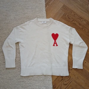Vit stickad tröja från Ami Paris - Vit stickad tröja från Ami Paris med röd hjärtlogga och bokstaven A på bröstet. Tröjan har rund halsringning, ribbade muddar och är långärmad. Perfekt för dig som gillar stilrena plagg med en twist.