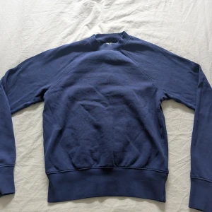 Mörkblå sweatshirt i bomull från Arket  - En basic mörkblå sweatshirt i mjuk bomull med rund hals och långa ärmar, från Arket. Tröjan har breda muddar vid ärmslut och nederkant, samt raglanärmar för en avslappnad vibe. Perfekt för dig som gillar enkel och stilren look.