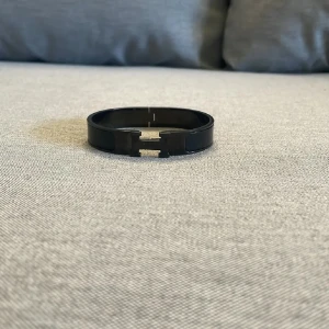 Svart  H-armband - Snyggt svart armband  med den ikoniska H-detaljen framtill. Armbandet är i metall med blank finish och har en stilren, modern look som passar perfekt till din outfit. Lätt att öppna och stänga med H-spännet.