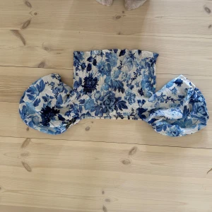 Blommig offshoulder topp från H&M - Supersöt offshoulder topp från H&M med blått blommönster på vit botten. Toppen har puffiga ärmar och smockad, elastisk överdel som sitter snyggt. Perfekt för dig som gillar romantisk och trendig stil. Materialet känns lätt och luftigt.