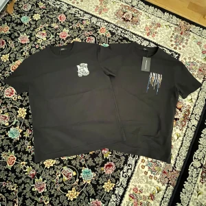 Amiri t shirts - 600 st. Två svarta t-shirts från Amiri i 100% bomull. Ena har färgstänk och stor AMIRI-logga på ryggen, den andra har en stor orm och AMIRI-tryck på ryggen samt mindre tryck fram. Klassisk passform och rund hals.