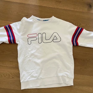 Vit sweatshirt från FILA med färgglada detaljer - Vit sweatshirt från FILA med stort logotryck framtill. Ärmarna har röda och blå ränder som ger en sportig känsla. Rund halsringning och mjukt material, perfekt för en avslappnad stil. Passar nog kvinnor men den är gjord för herr.